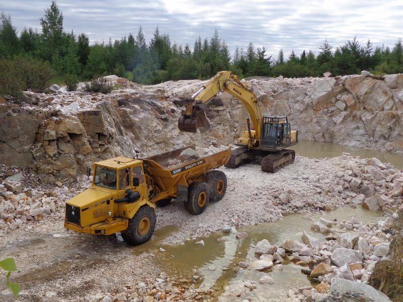 A digger and dump truck work to load up some natural stone in a small quarry.