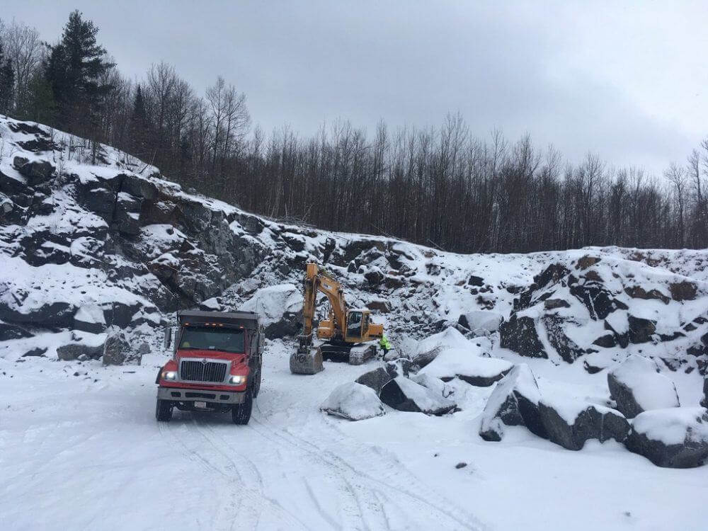 A snow-covered quarry is shown with a digger and dump truck in the foreground.