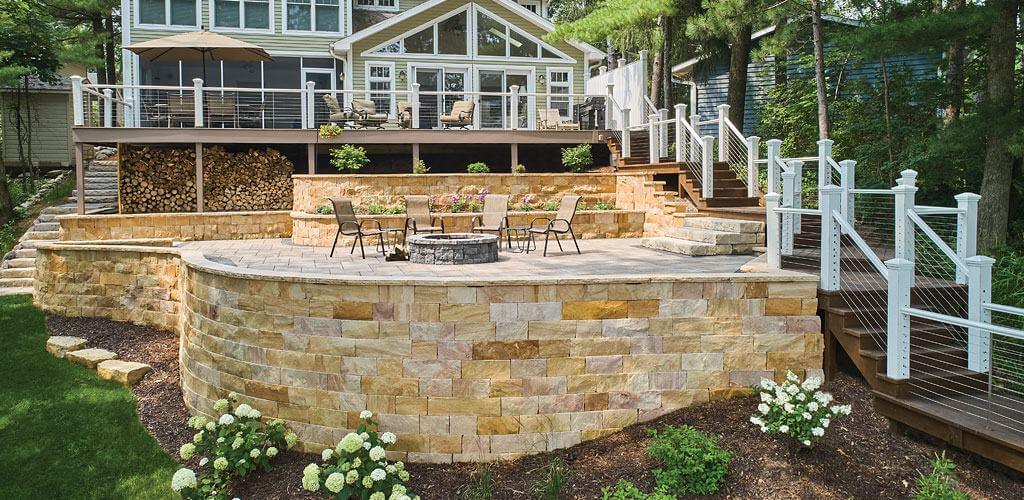 A beautiful stone retaining wall creates an elevated patio for a bonfire pit in a sloping backyard.